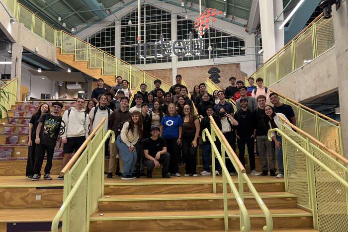 Cerca de quarenta e cinco participantes do bootcamp posando na escadaria central do Inteli, com o letreiro da faculdade na parede ao fundo.