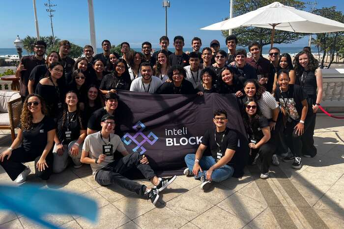Cerca de quarenta membros do Inteli Blockchain reunidos ao ar livre à beira-mar, de camiseta do clube e crachá, segurando a bandeira do clube.