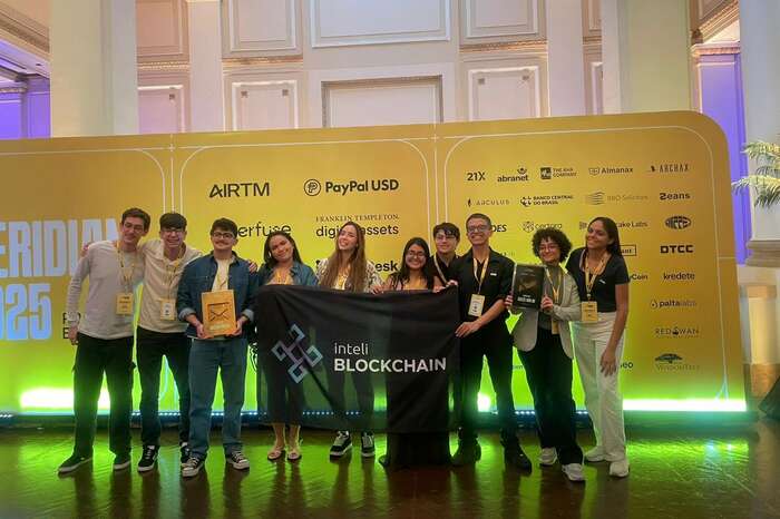 Dez membros do clube no palco do Meridian 2025 segurando a bandeira do Inteli Blockchain, dois deles com os troféus nas mãos, diante do painel de patrocinadores do evento.