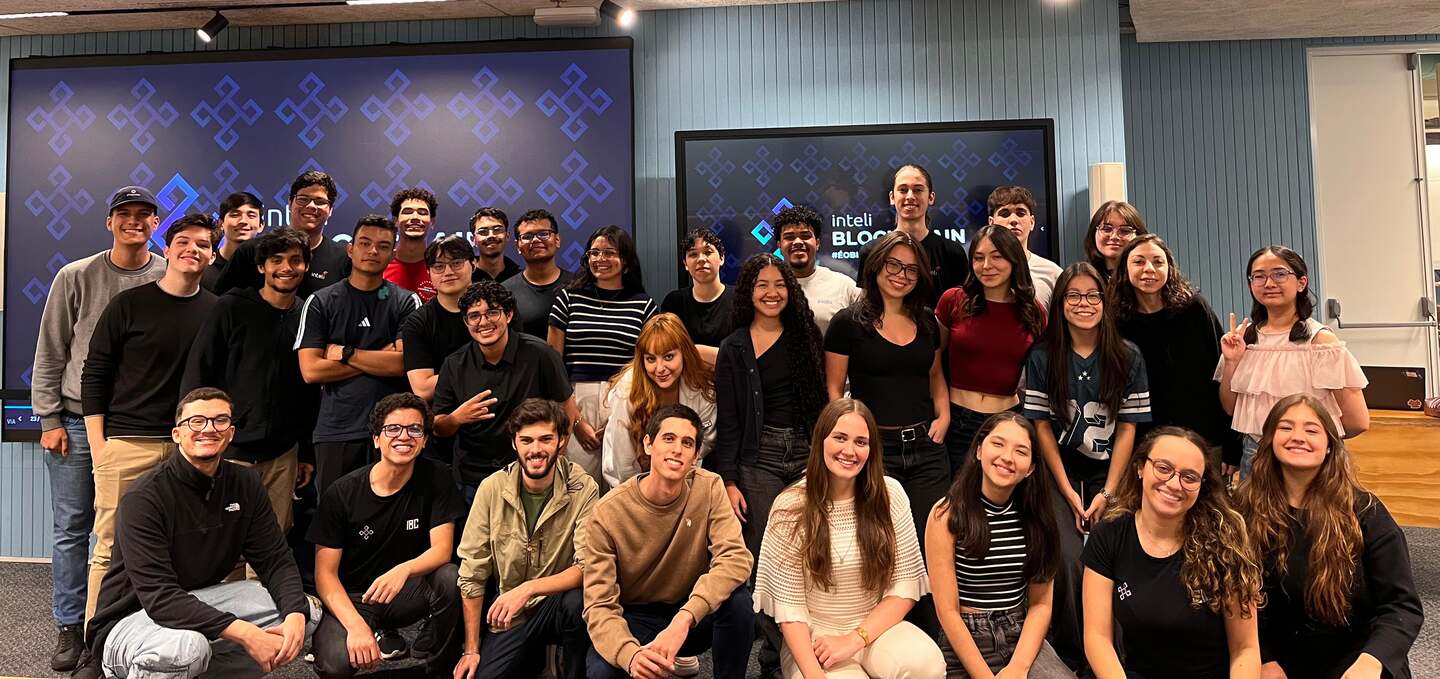 Cerca de trinta e cinco membros do Inteli Blockchain posando juntos em duas fileiras num auditório do Inteli, com os telões atrás exibindo a marca do clube.