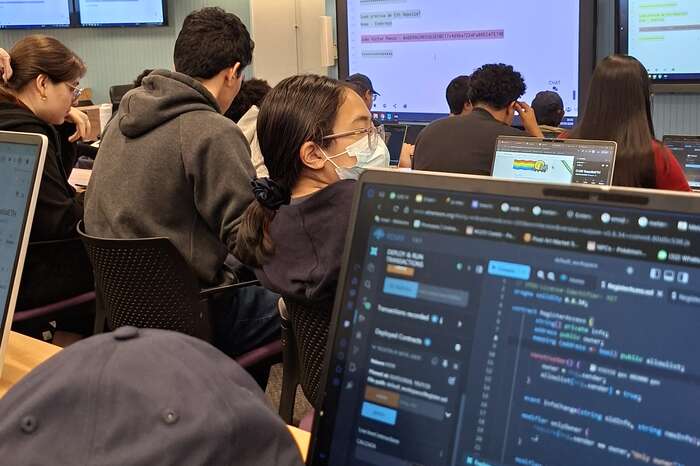 Sala do Inteli durante o workshop: alunos em fileiras com notebooks abertos, telas de projeção ao fundo e, em primeiro plano, um notebook com código de contrato aberto no editor.